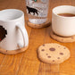 Two mugs with pixelated designs on cork coasters that look like chocolate chip cookies on a wooden surface.