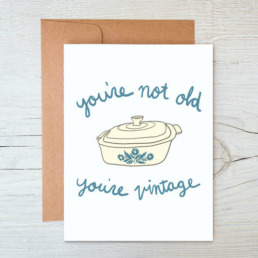 Greeting card with a vintage corningware pot illustration and text "you're not old you're vintage" on a wooden surface
