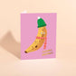 Greeting card with a banana wearing a green hat and striped scarf on a pink background