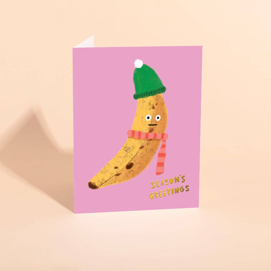 Greeting card with a banana wearing a green hat and striped scarf on a pink background