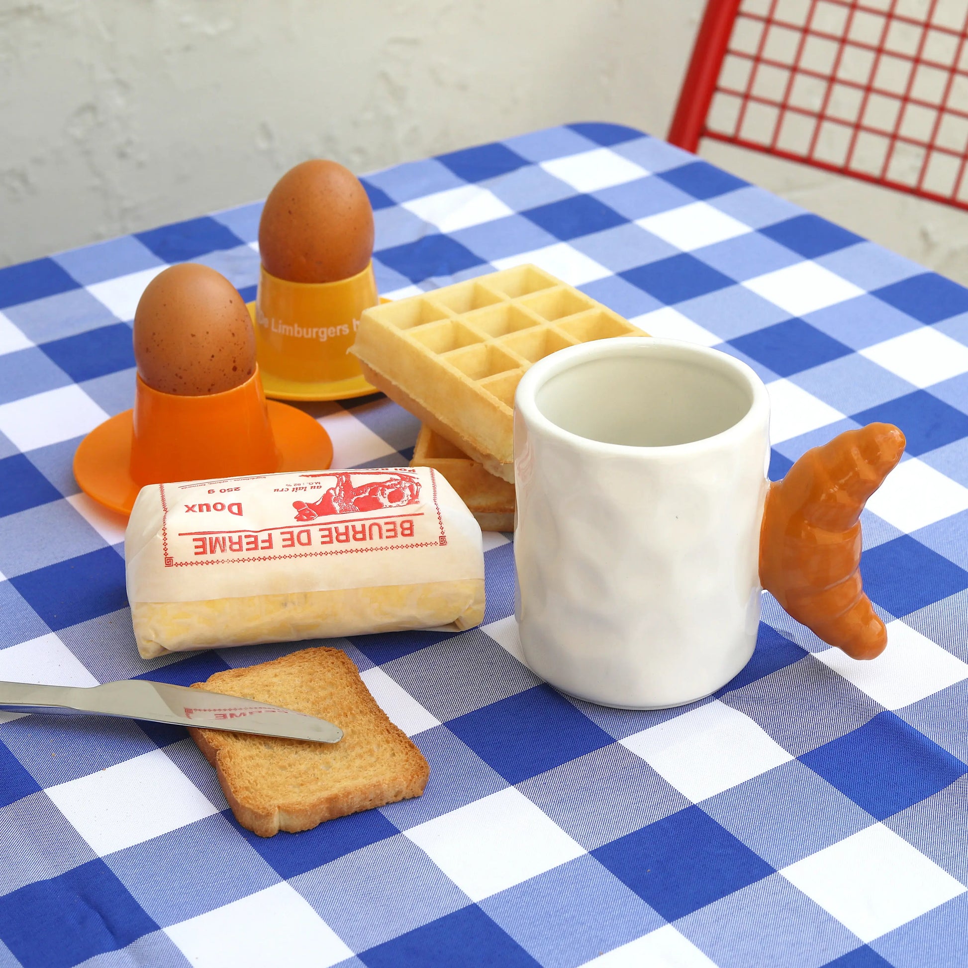 white, ceramic mug with a brown handle shaped like a croissant. Photographed with other breakfast items including eggs, toast, waffles and butter. 