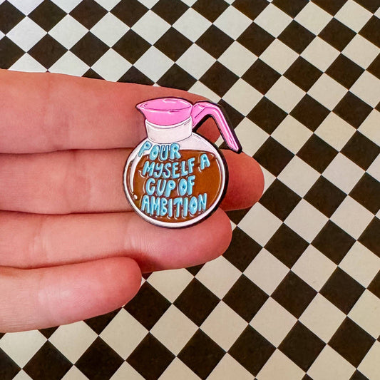 Enamel pin of a coffee pot with 'Pour myself a cup of ambition' text held by a hand on a checkered background