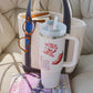 A white handled tumbler with a straw sits on top of a purple and blue book, in front of a cream tote bag with navy trim. The tumbler has red text and chili pepper graphics.