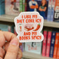 A hand holds a die-cut sticker featuring the text 'I LIKE MY DIET COKE ICY AND MY BOOKS SPICY' with illustrations of a Diet Coke can, books, and a red chili pepper, set against a blurred background of bookshelves.