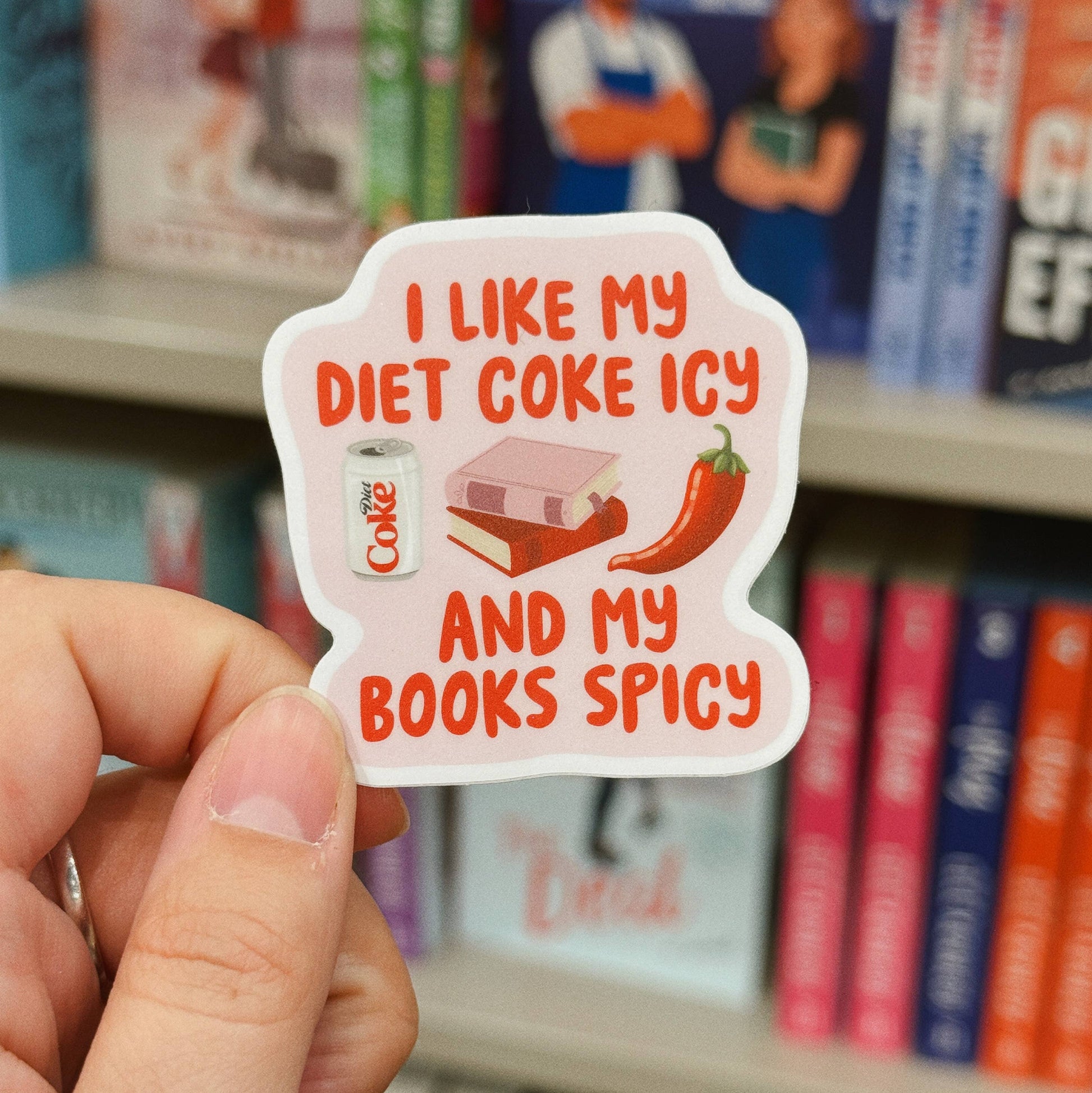 A hand holds a die-cut sticker featuring the text 'I LIKE MY DIET COKE ICY AND MY BOOKS SPICY' with illustrations of a Diet Coke can, books, and a red chili pepper, set against a blurred background of bookshelves.