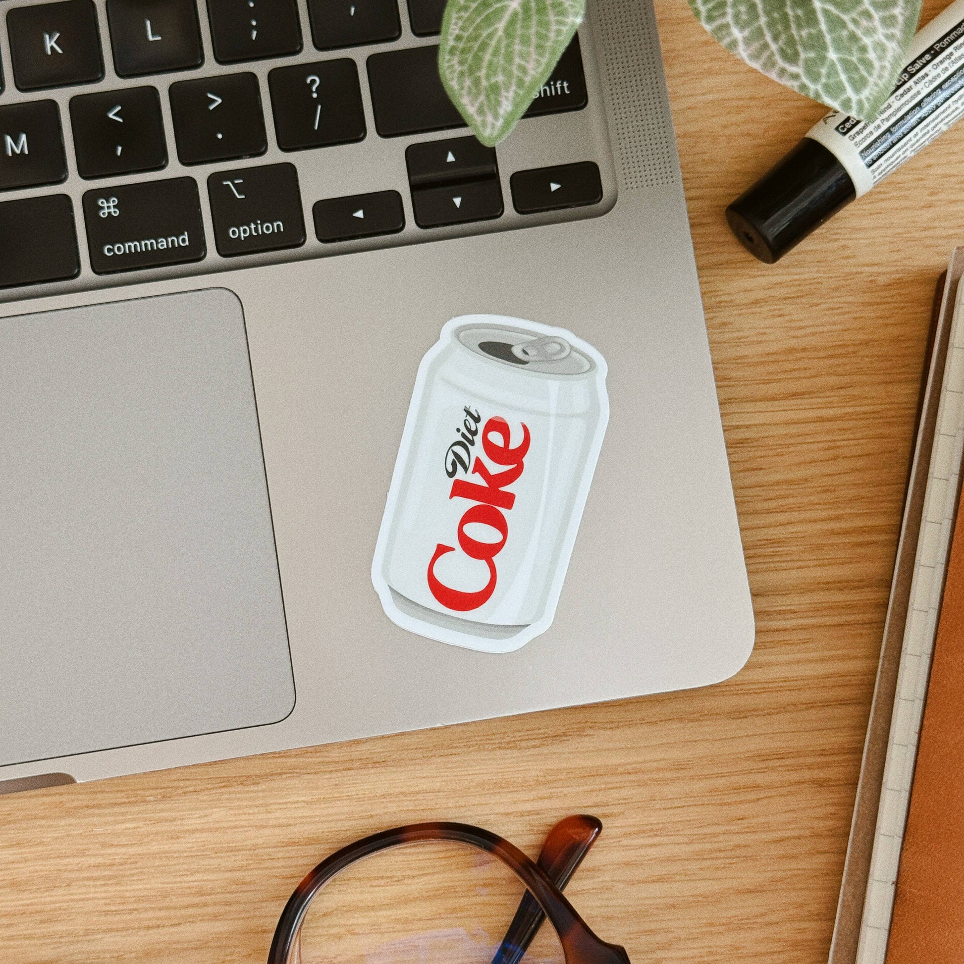 A die-cut sticker of a white Diet Coke can with red lettering, shown applied to the palm rest of a silver laptop on a wooden desk, with glasses, a marker, and a plant nearby.