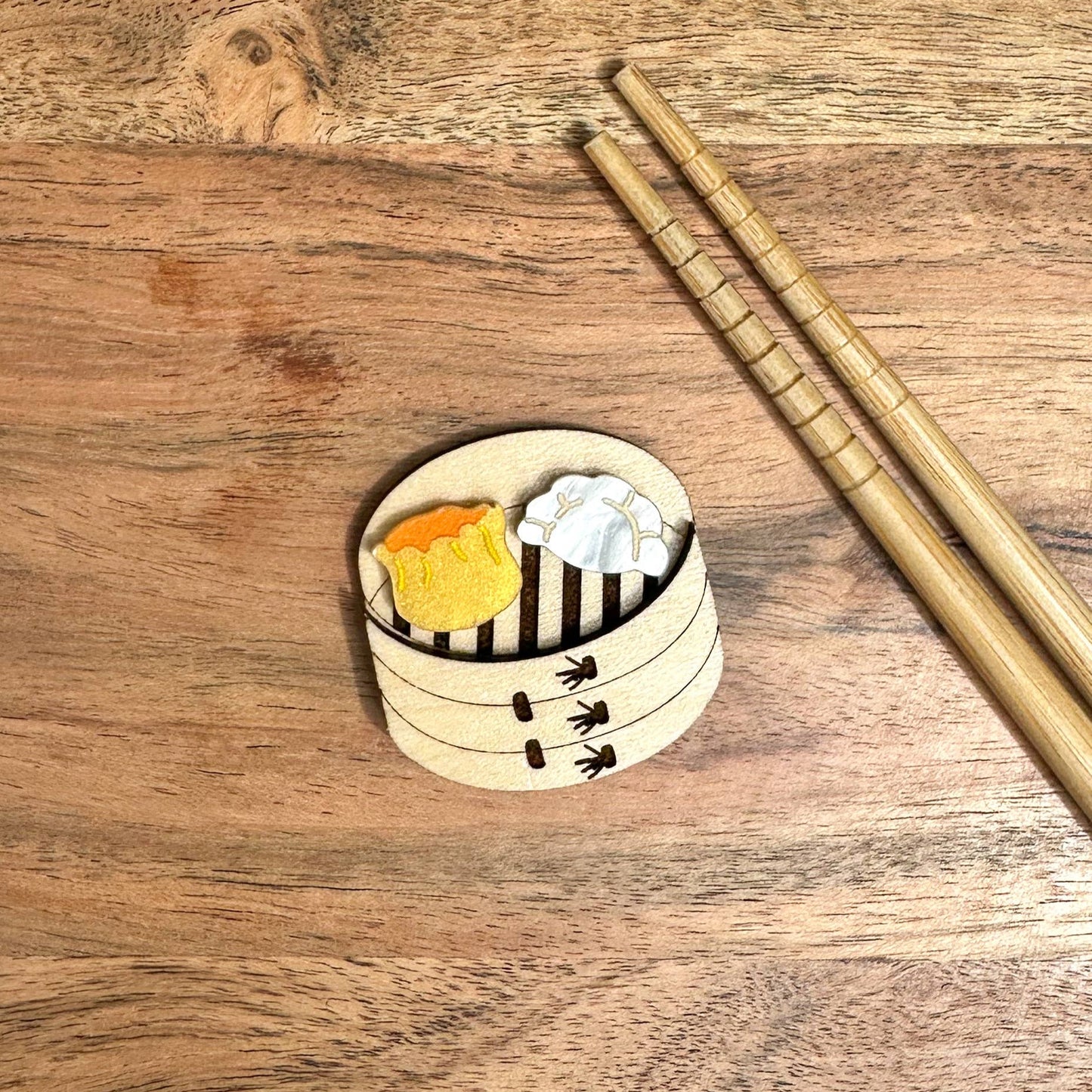 A brooch/lapel pin of a bamboo steamer with a siu mai and dumpling in it