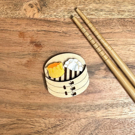 A brooch/lapel pin of a bamboo steamer with a siu mai and dumpling in it