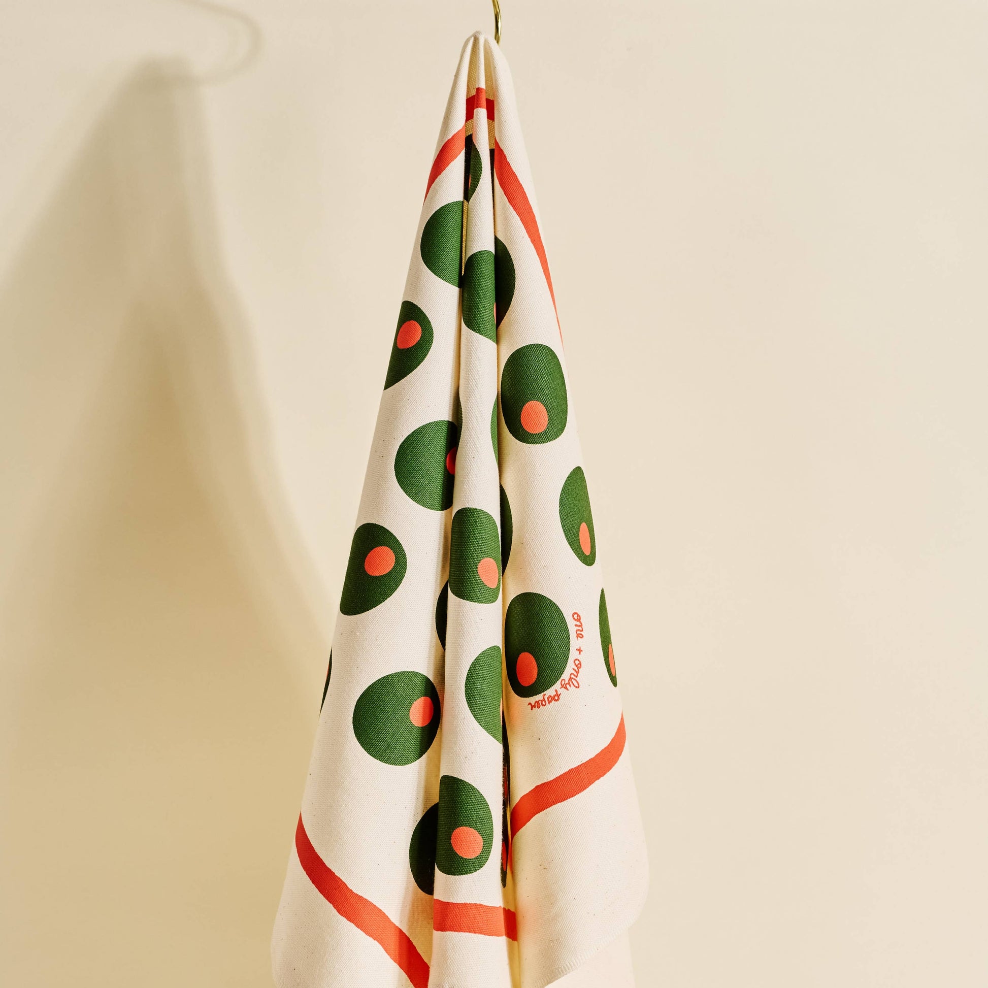 Decorative towel with pimento stuffed green olives pattern, hanging on a hook against a beige wall.