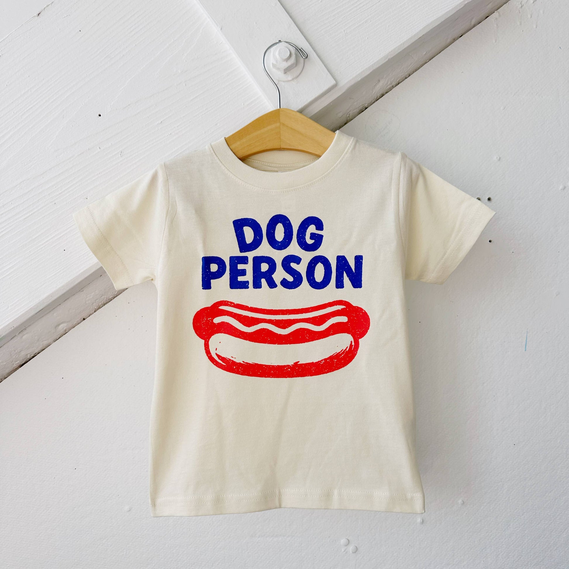 White t-shirt with 'Dog Person' text and hot dog graphic on a wooden hanger against a white background