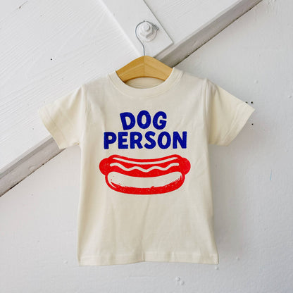White t-shirt with 'Dog Person' text and hot dog graphic on a wooden hanger against a white background