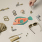 Collection of small items including a fish-shaped pouch, sunglasses, and cosmetic products on a beige surface.