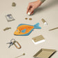 Hand interacting with a fish-shaped pouch on a beige surface with various items around
