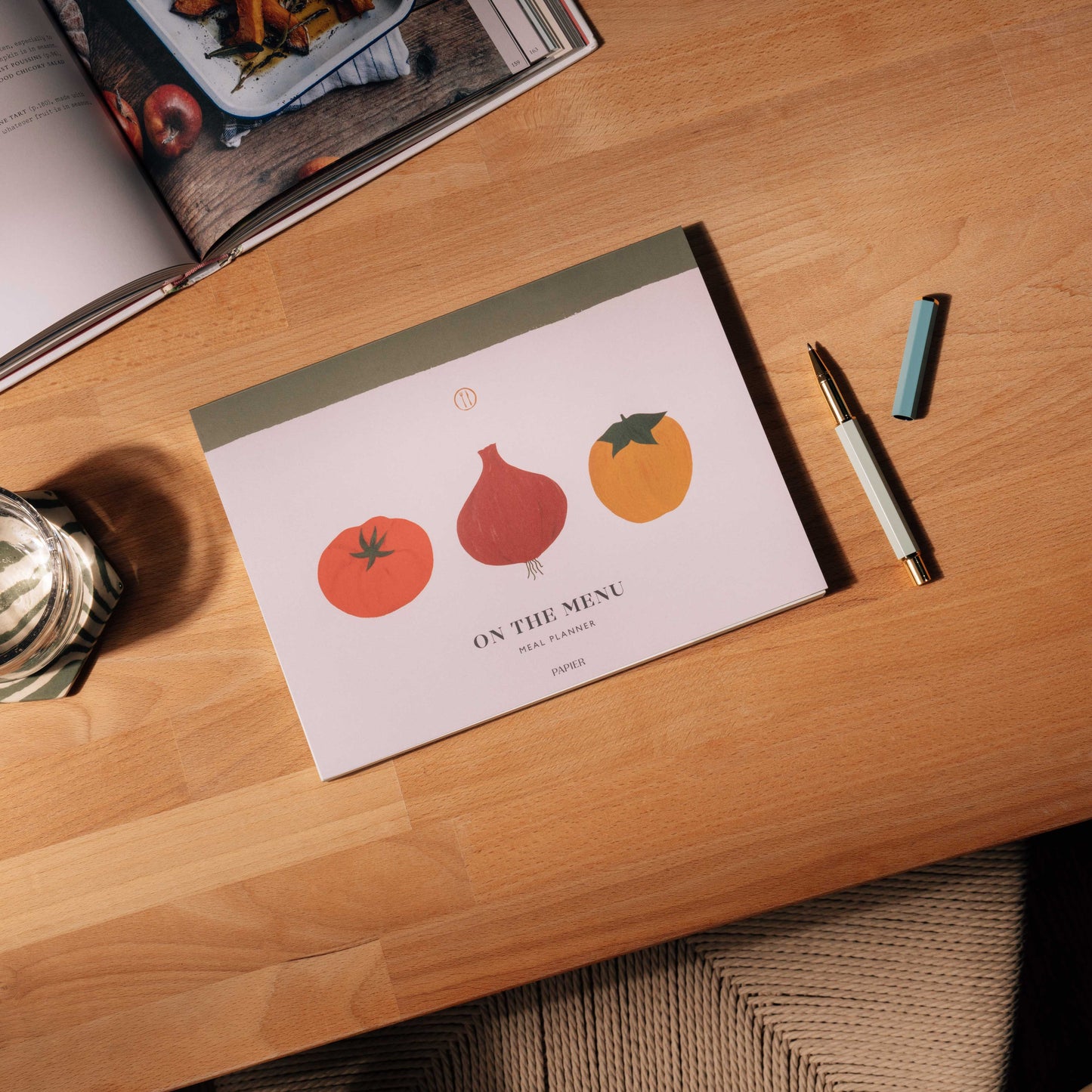 Front cover of Vegetable Medley Meal Planner. Features illustration of tomato, red onion and yellow apple on neutral background with rustic green stripe along the spine. Cover text reads "ON THE MENU... meal planner. " Shown on wood table with recipe bookand pen.