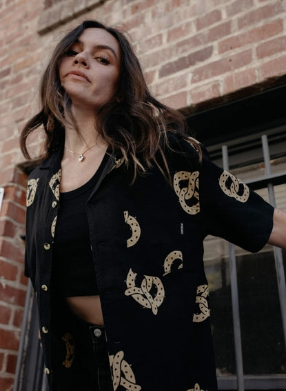 Photo of woman with long hair wearing pretzel shirt unbuttoned over black tank top, with black jeans.