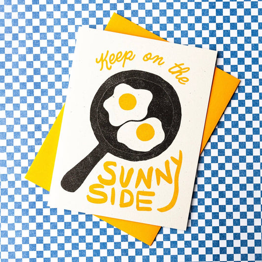 Greeting card with two sunny side up eggs in a skillet with text that reads "Keep on the sunny side". Background of image is blue and white checkerboard 