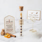 A glass tube wand candle with a cork stopper and gold lettering stands on a marble surface beside printed cards and small props. The product is a clear glass tube containing a striped candle, with a cork stopper and a gold label reading 'CANDY CANDLE.