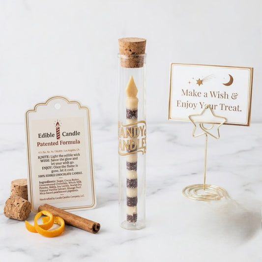 A glass tube wand candle with a cork stopper and gold lettering stands on a marble surface beside printed cards and small props. The product is a clear glass tube containing a striped candle, with a cork stopper and a gold label reading 'CANDY CANDLE.