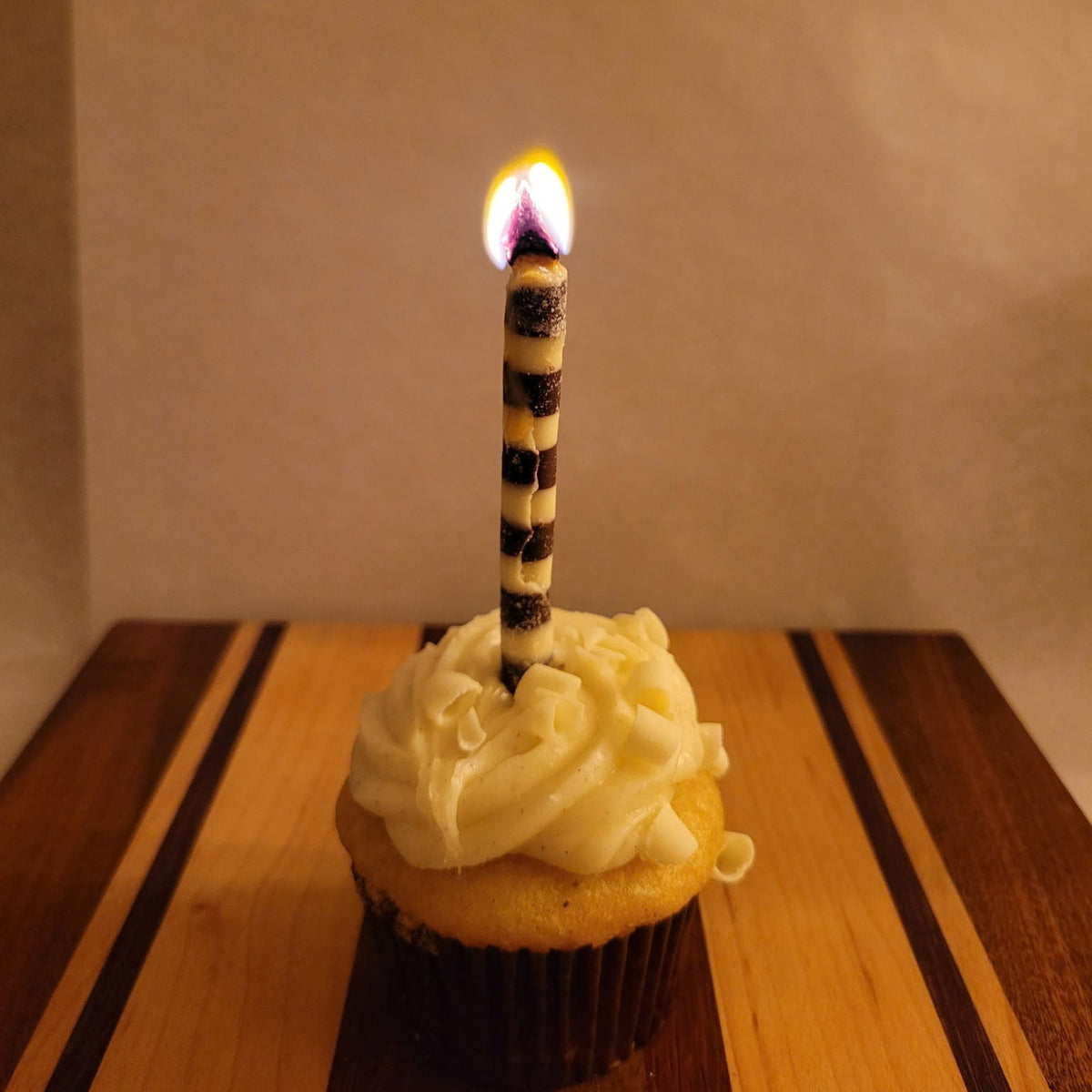 A single birthday candle with a black and white striped pattern is lit and placed in the center of a frosted cupcake on a wooden board against a plain beige background.