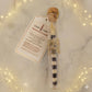 A glass tube candle with a cork stopper and gold seal, containing a black-and-white striped wax candle, shown with a printed product tag on a light marble surface with a sparkling overlay.