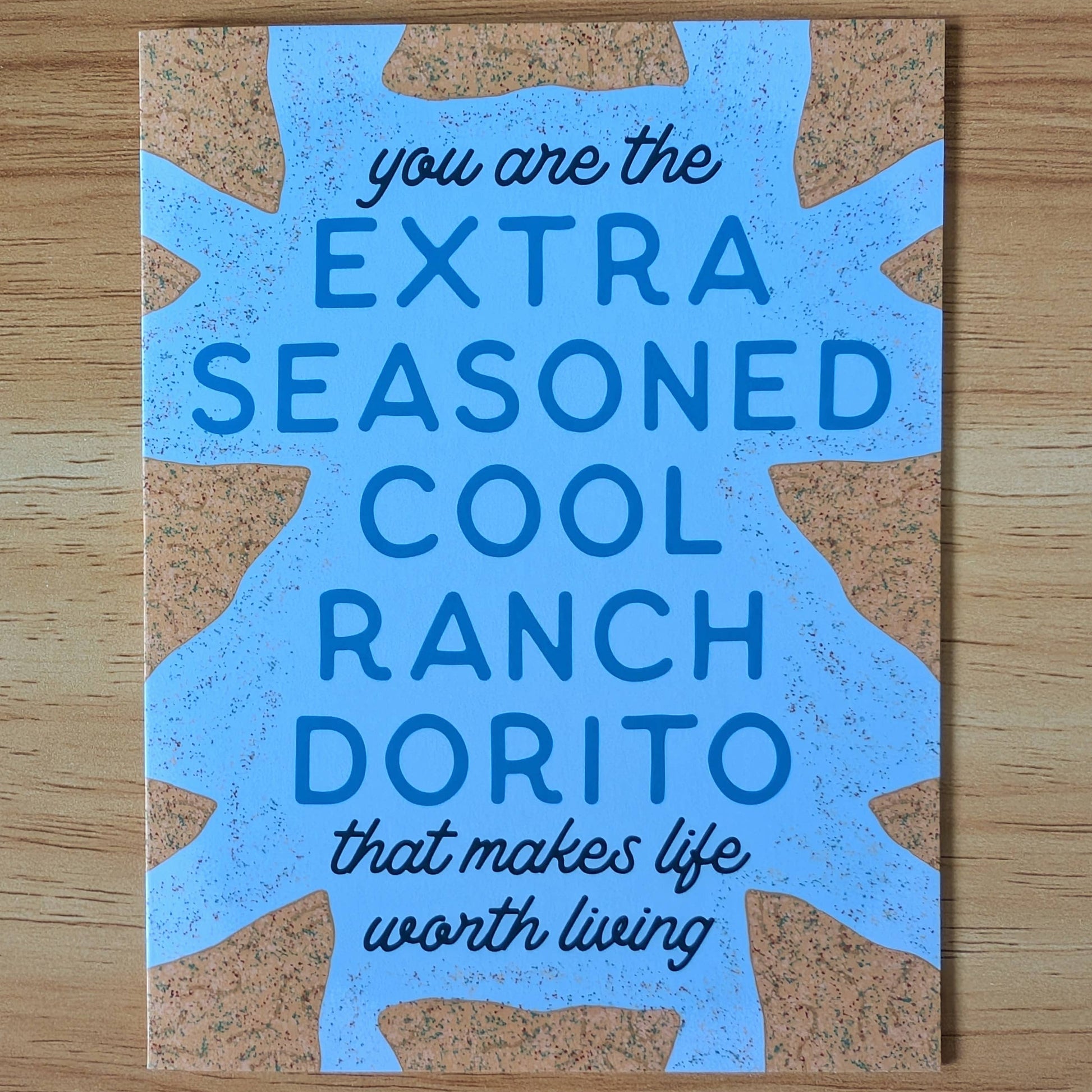 Card with blue text featuring a humorous message, "you are the extra seasoned cool ranch dorito that makes life worth living" on a wooden surface.