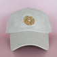 Khaki colored cap with a embroidered everything bagel design on front, pictured here on a pink background
