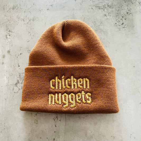 Dark brown knit beanie hat with "chicken nuggets" embroidered in light brown font.