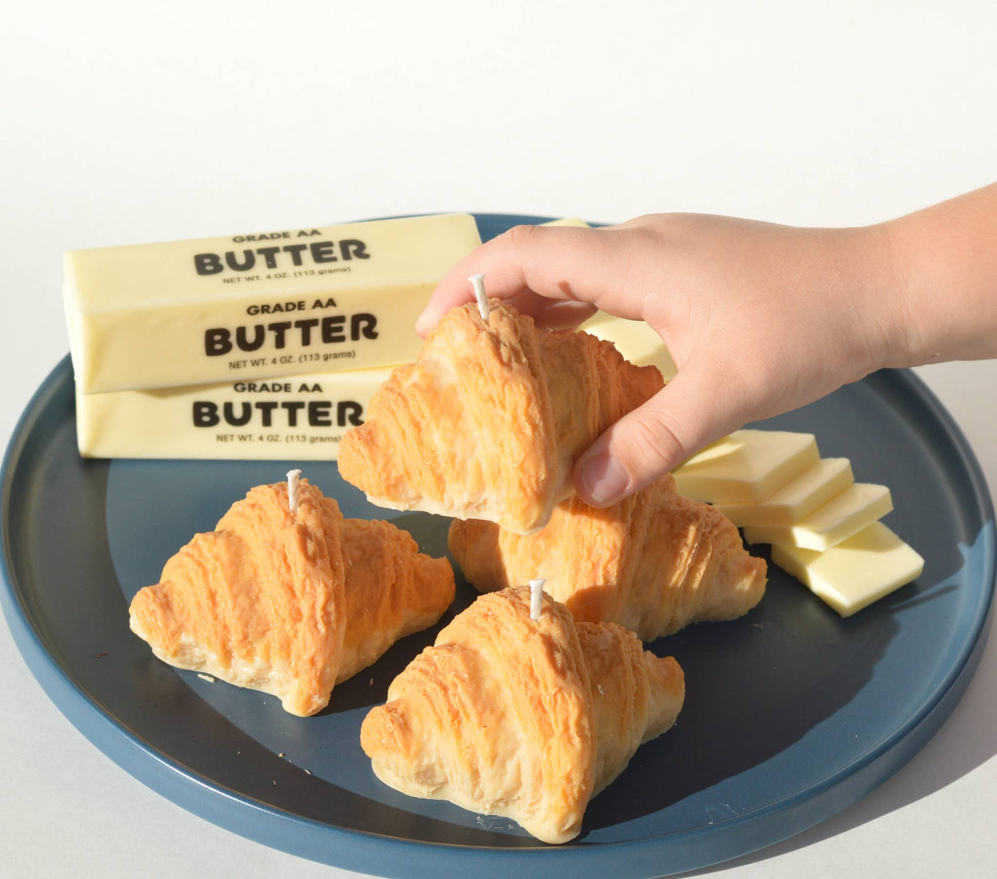Hand reaching for a pile of four croissant candles on a plate next to slabs of butter.