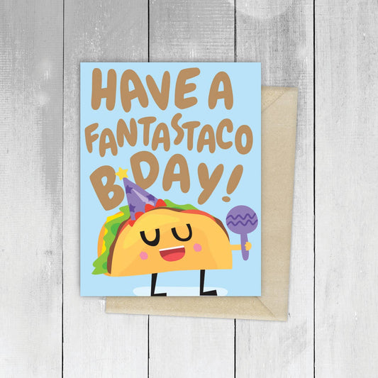 Birthday card with a cartoon taco and text 'Have a fantastaco bday!' on a wooden surface.