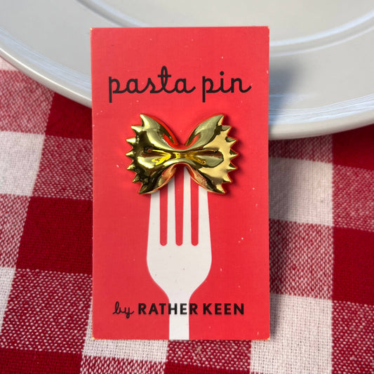 Gold pasta-shaped pin on a red card with 'RATHER KEEN' branding, placed on a red and white checkered tablecloth.