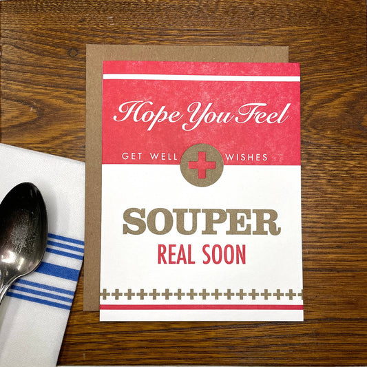 Get well card with 'Hope You Feel Souper Real Soon' text on a wooden surface with a spoon and towel.