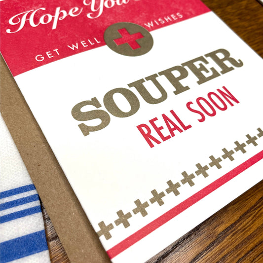Get well card with 'Hope you feel Souper Real Soon' text on a wooden surface