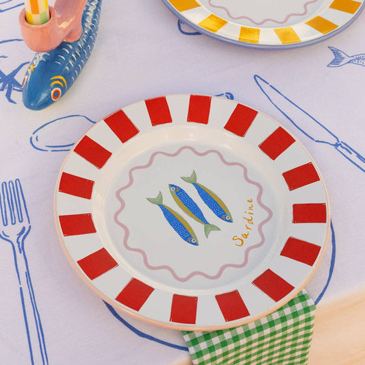 Enamel plate with red and white striped rim, fish design with cutlery and a checkered cloth.