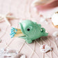 A mint green ceramic fish-shaped match holder containing blue-tipped matches, styled on a pink surface with seashells, netting, and clear stones.