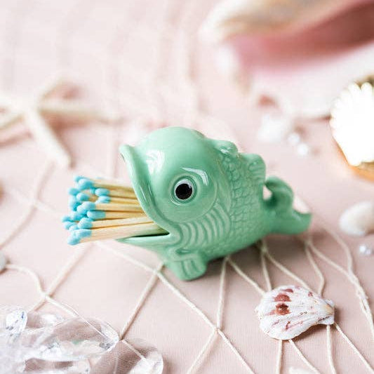 A mint green ceramic fish-shaped match holder containing blue-tipped matches, styled on a pink surface with seashells, netting, and clear stones.