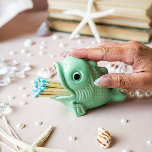Hand holding a green fish-shaped matchbox with matches on a decorative surface with starfish and books.