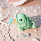A small mint green fish-shaped match holder holding blue and yellow matches, styled on a beige surface with shells, starfish, and pearl-like beads.