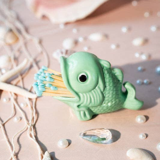 A small mint green fish-shaped match holder holding blue and yellow matches, styled on a beige surface with shells, starfish, and pearl-like beads.