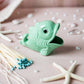 Green fish-shaped match holder with matches on a pink surface with starfish and pearls.