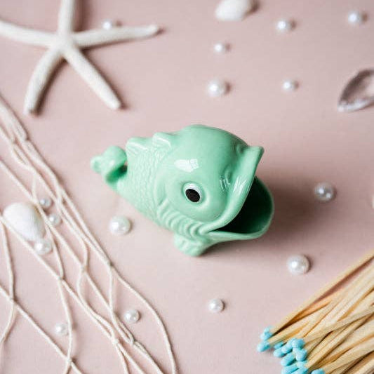 A small mint green fish-shaped match holder with an open mouth, styled on a dusty pink background with seashell props, pearl beads, netting, and matchsticks.