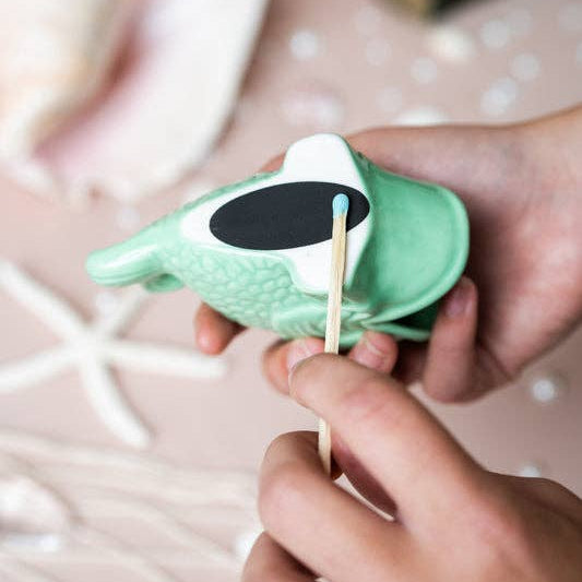 A close-up of hands holding a small mint green seashell-shaped item with a black oval patch and white tip, using a wooden stick to apply or press material onto it, set against a soft pink background with blurred seashell props.