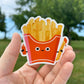 Sticker of a cute cartoon-style French fry container held in a hand with a natural background.