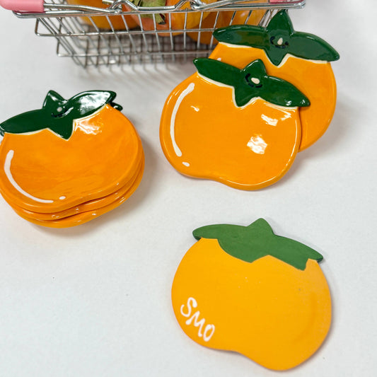 Set of persimmon trinket dishes with green leaves on a white surface, with a small basket of persimmons in the background.
