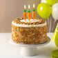 Carrot cake with carrot-shaped candles on a white cake stand, surrounded by balloons.