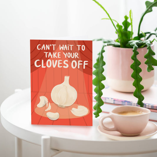 red greeting card with a garlic bulb centerstage, under a spotlight with text above that reads "can't wait to take your cloves off" 