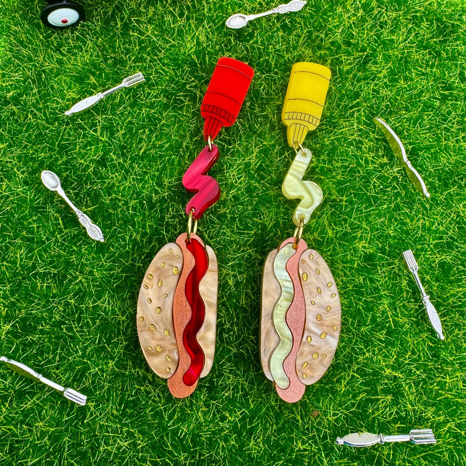 Hot dog earrings with ketchup and mustard details on them. Earrings are pictured on grass with a toy wagon and utensils.