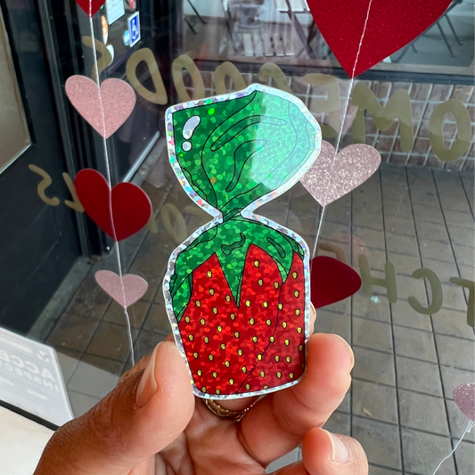 Hand holding a colorful strawberry-shaped candy sticker with hearts in the background.