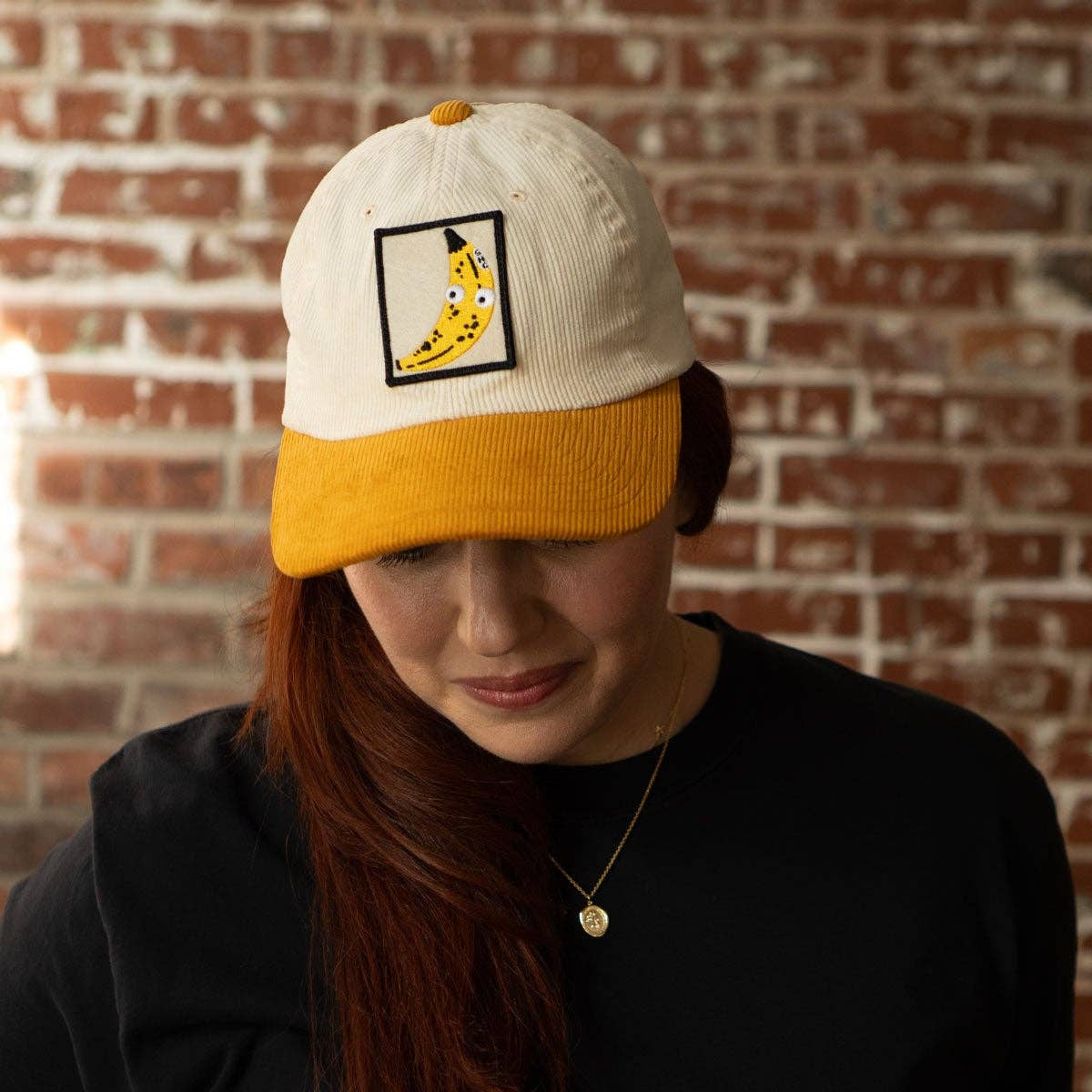 Person wearing a cap with a banana graphic patch against a brick wall