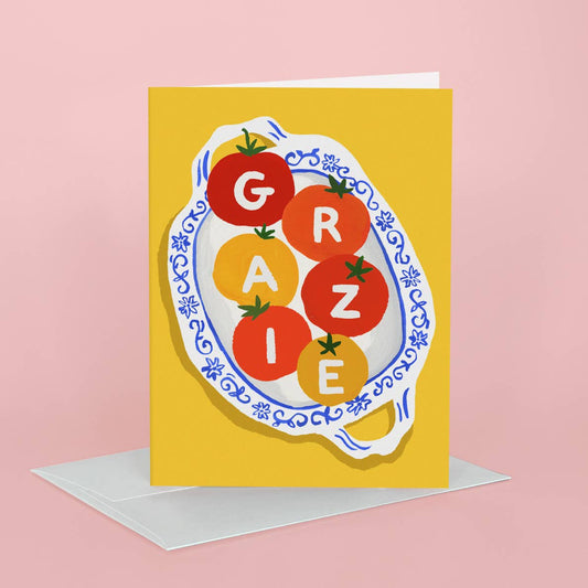 A yellow greeting card with a stylized tomato illustration spelling 'GRAZIE' on a plate, shown standing upright with a white envelope against a pink background.