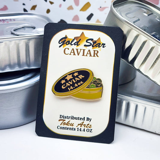 Gold Star Caviar labeled card packaging with the green caviar enamel pin on it, set against plain tinned fish cans in the background